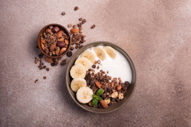 Yoğurtlu granola, taze muz, çikolata damlaları ve badem fıstıklı sağlıklı kahvaltı için kahverengi beton arka planda