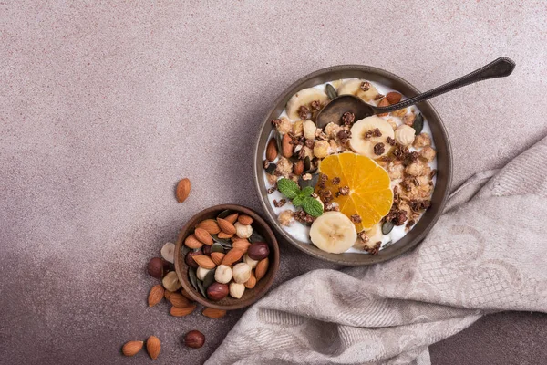 Yoğurtlu granola, taze muz, portakal, badem ve fındık, kahverengi beton zemin üzerinde sağlıklı kahvaltı için kabak çekirdeği.