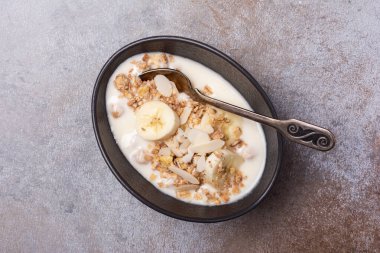 Sağlıklı kahvaltı için yoğurtlu granola, taze muz ve badem fıstığı gri beton arka planda.