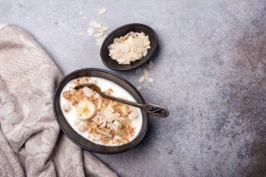 Sağlıklı kahvaltı için yoğurtlu granola, taze muz ve badem fıstığı gri beton arka planda.