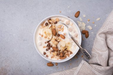 Sağlıklı kahvaltı için yoğurtlu granola, taze muz ve badem fıstığı gri beton arka planda.