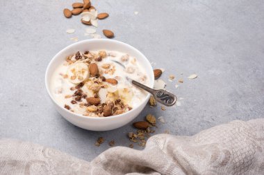 Sağlıklı kahvaltı için yoğurtlu granola, taze muz ve badem fıstığı gri beton arka planda.