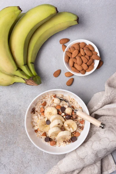 Taze muz dilimli yulaf ezmesi, badem, fındık ve çikolata, sağlıklı kahvaltı için gri beton arka planda.