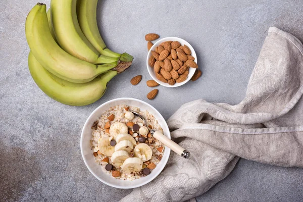 Taze muz dilimli yulaf ezmesi, badem, fındık ve çikolata, sağlıklı kahvaltı için gri beton arka planda.