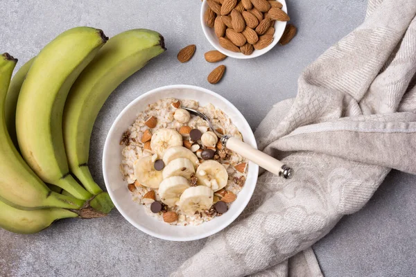 Taze muz dilimli yulaf ezmesi, badem, fındık ve çikolata, sağlıklı kahvaltı için gri beton arka planda.