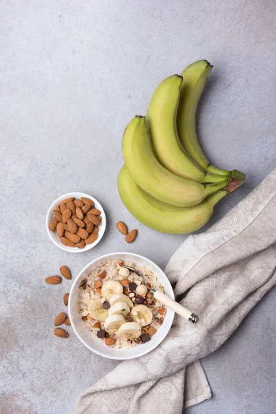 Taze muz dilimli yulaf ezmesi, badem, fındık ve çikolata, sağlıklı kahvaltı için gri beton arka planda.