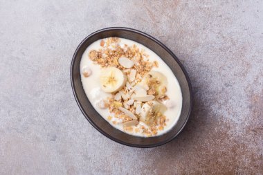 Sağlıklı kahvaltı için yoğurtlu granola, taze muz ve badem fıstığı gri beton arka planda.