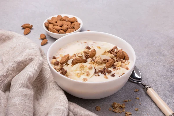 Sağlıklı kahvaltı için yoğurtlu granola, taze muz ve badem fıstığı gri beton arka planda.
