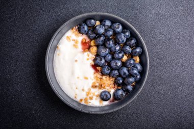 Top view with bowl of healthy dessert or breakfast with yogurt, blueberries and granola on black concrete background