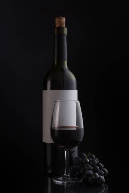 Bottle of red wine with white template or mock-up label, glass and fresh grape on black background