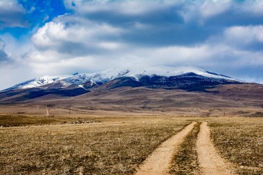 Toprak bir yol dağa çıkıyor. Dağın üstünde beyaz bulutlar. Dağın tepesi karla kaplı.