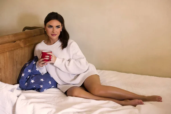 Çekici Genç Bir Kadın Beyaz Bir Kazakla Yatakta Bir Kupa — Stok fotoğraf
