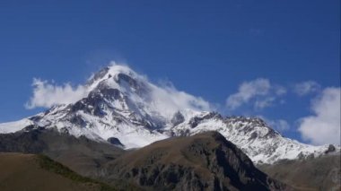 Kazbek Dağı ya da Kazbegi Dağı karla kaplı. Rüzgar dağın tepesinden kar esiyor. Stepantsminda, Georgia.