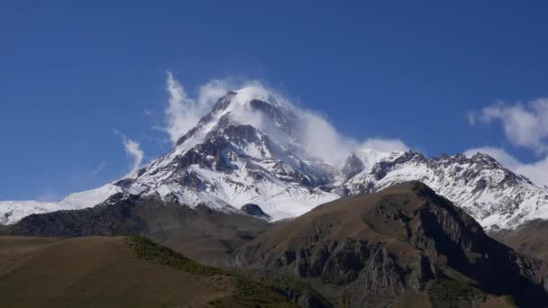 Der Berg Kazbek oder Kazbegi ist mit Schnee bedeckt. Der Wind bläst Schnee vom Gipfel des Berges. Stepantsminda, Georgien. — Stockvideo