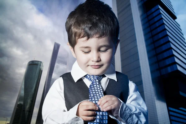 Child dressed businessman with hands in his tie and skyscrapers — Stock Photo, Image