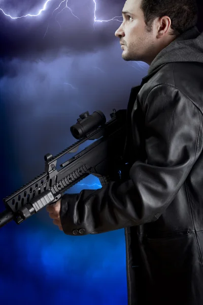 Hombre con chaqueta de cuero largo y rifle de asalto sobre tormenta backgr —  Fotos de Stock