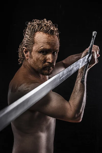 Strong warrior licking a big sword, covered in mud and naked — Stock Photo, Image