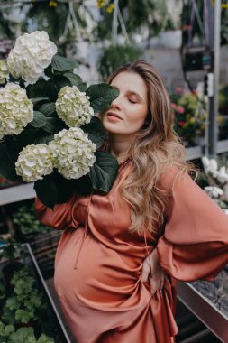 Genç hamile bir kadın bir serada çiçekleri suluyor. Evde ilkbahar ve pencerenin dışında kış