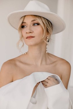 Kendine güvenen genç sarışın kadın gülümsüyor, beyaz arka planda izole edilmiş kameraya bakıyor. Beyaz takım elbiseli, beyaz şapkalı, beyaz duvarda poz veren başarılı bir kadının stüdyo portresi..