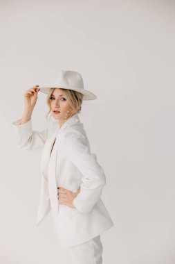 Kendine güvenen genç sarışın kadın gülümsüyor, beyaz arka planda izole edilmiş kameraya bakıyor. Beyaz takım elbiseli, beyaz şapkalı, beyaz duvarda poz veren başarılı bir kadının stüdyo portresi..