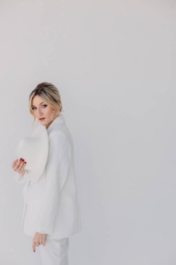 Kendine güvenen genç sarışın kadın gülümsüyor, beyaz arka planda izole edilmiş kameraya bakıyor. Beyaz takım elbiseli, beyaz şapkalı, beyaz duvarda poz veren başarılı bir kadının stüdyo portresi..