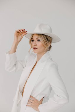 Kendine güvenen genç sarışın kadın gülümsüyor, beyaz arka planda izole edilmiş kameraya bakıyor. Beyaz takım elbiseli, beyaz şapkalı, beyaz duvarda poz veren başarılı bir kadının stüdyo portresi..