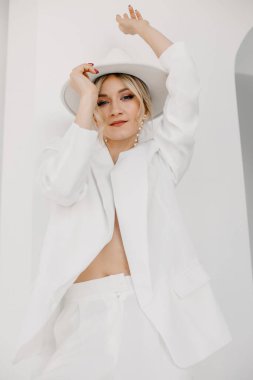 Confident young blond woman smiling, looking at camera isolated on white background. Studio portrait of successful friendly female in white suit and hat, posing over white wall.