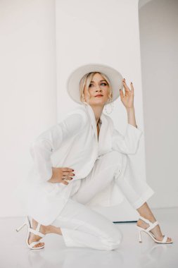 Kendine güvenen genç sarışın kadın gülümsüyor, beyaz arka planda izole edilmiş kameraya bakıyor. Beyaz takım elbiseli, beyaz şapkalı, beyaz duvarda poz veren başarılı bir kadının stüdyo portresi..