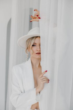 Kendine güvenen genç sarışın kadın gülümsüyor, beyaz arka planda izole edilmiş kameraya bakıyor. Beyaz takım elbiseli, beyaz şapkalı, beyaz duvarda poz veren başarılı bir kadının stüdyo portresi..