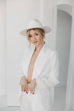 Kendine güvenen genç sarışın kadın gülümsüyor, beyaz arka planda izole edilmiş kameraya bakıyor. Beyaz takım elbiseli, beyaz şapkalı, beyaz duvarda poz veren başarılı bir kadının stüdyo portresi..