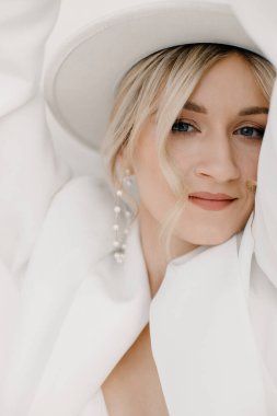 Kendine güvenen genç sarışın kadın gülümsüyor, beyaz arka planda izole edilmiş kameraya bakıyor. Beyaz takım elbiseli, beyaz şapkalı, beyaz duvarda poz veren başarılı bir kadının stüdyo portresi..