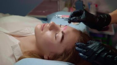 Usta kaş makyajı prosedürünü kuaförde kadına özel iğne dövme makinesiyle yapıyor. Mikropatenli kaş dövmesi. Koyu pigmentler derinin altına enjekte edilir. Kozmetoloji prosedürü