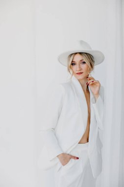 Kendine güvenen genç sarışın kadın gülümsüyor, beyaz arka planda izole edilmiş kameraya bakıyor. Beyaz takım elbiseli, beyaz şapkalı, beyaz duvarda poz veren başarılı bir kadının stüdyo portresi..