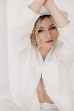 Kendine güvenen genç sarışın kadın gülümsüyor, beyaz arka planda izole edilmiş kameraya bakıyor. Beyaz takım elbiseli, beyaz şapkalı, beyaz duvarda poz veren başarılı bir kadının stüdyo portresi..