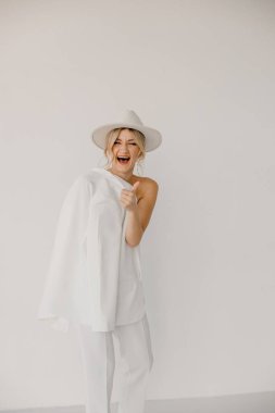 Kendine güvenen genç sarışın kadın gülümsüyor, beyaz arka planda izole edilmiş kameraya bakıyor. Beyaz takım elbiseli, beyaz şapkalı, beyaz duvarda poz veren başarılı bir kadının stüdyo portresi..