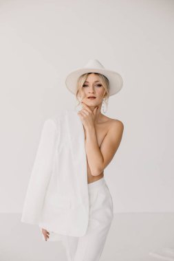 Kendine güvenen genç sarışın kadın gülümsüyor, beyaz arka planda izole edilmiş kameraya bakıyor. Beyaz takım elbiseli, beyaz şapkalı, beyaz duvarda poz veren başarılı bir kadının stüdyo portresi..