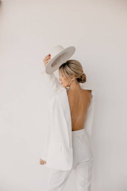 Kendine güvenen genç sarışın kadın gülümsüyor, beyaz arka planda izole edilmiş kameraya bakıyor. Beyaz takım elbiseli, beyaz şapkalı, beyaz duvarda poz veren başarılı bir kadının stüdyo portresi..