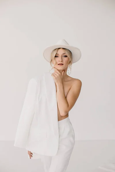 Confident young blond woman smiling, looking at camera isolated on white background. Studio portrait of successful friendly female in white suit and hat, posing over white wall. — Stockfoto