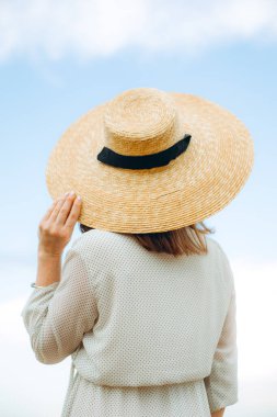 Baharda öğleden sonra mavi kurdeleli hasır şapkalı bir kız. Arkadan bak. Havalı günlük yaz ya da bahar kıyafeti. Kayıkçı şapkalı bir kadın. Kadın bahar modası konsepti. Çiçekli leylak çalıları