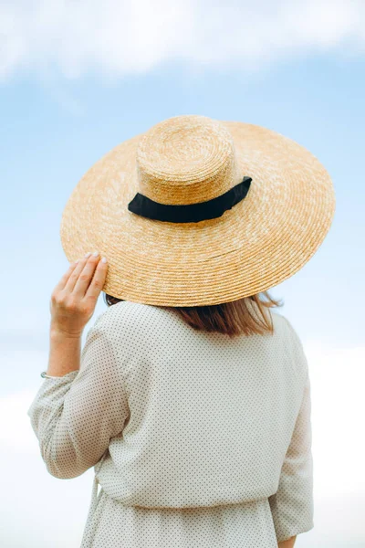 Girl in a straw hat with a blue ribbon on a spring afternoon. Back view. Trendy casual summer or spring outfit. Woman in a boatmans straw hat. concept of female spring fashion. Flowering lilac bushes