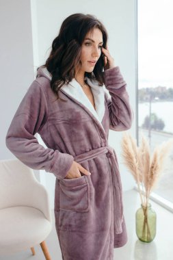 Beyaz bornozlu bir kızın büyük pencereli bir odada küvetin yanında durduğunu hayal et. Yüksek kalite fotoğraf