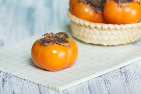 Three persimmons on the table. One on the napkin and two in the basket — Stock Photo, Image