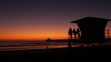 Cankurtaran kulesinin yanındaki genç kızların siluetleri Pasifik Okyanusu sahilindeki arkadaşları Oceanside, Kaliforniya 'da günbatımı. Tanınmayan gençler, insanlar ve alacakaranlık mor gökyüzü.