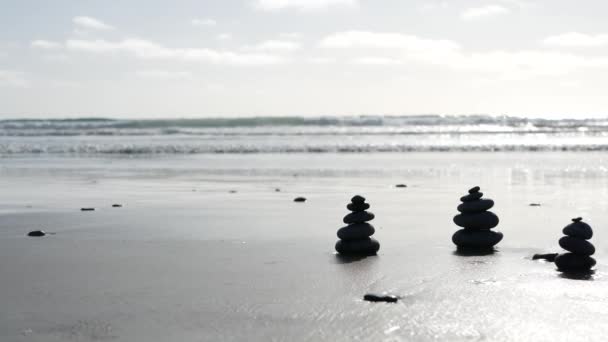 Szikla egyensúlyozik az óceán partján, kövek halmozódnak fel a tengervíz hullámainál. Kavicsok piramisa a homokos parton — Stock videók
