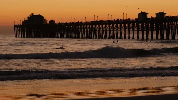 Sylwetka pomostu o zachodzie słońca, Kalifornia, USA, Oceanside. Ośrodek surfingowy, tropikalna plaża oceanu. Surfer czeka na falę. — Zdjęcie stockowe