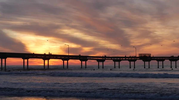 Surfistas a surfar por cais. Ondas de água do oceano, pessoas e céu ao pôr-do-sol. Califórnia — Fotografia de Stock