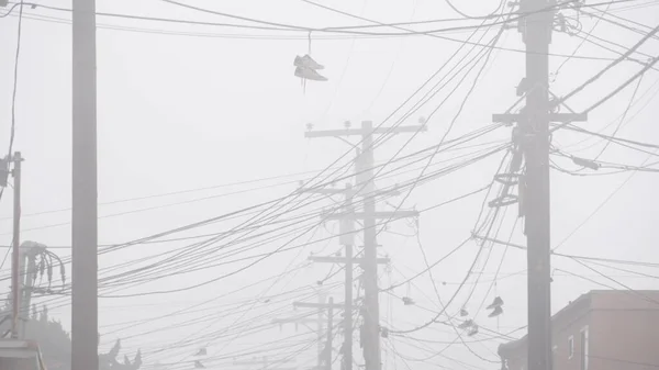 Sneakers canvas shoes hanging on power line, fog on city street. Boots on wires. — Stock Photo, Image