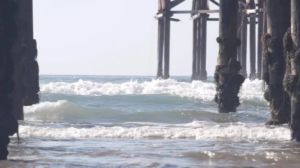 Debajo del muelle de cristal de madera en pilas, olas de agua de la playa del océano, California EE.UU.. — Foto de Stock
