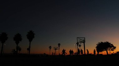 Basketbol sahasındaki oyuncuların siluetleri, basketbol oynayan insanlar, günbatımı okyanus plajı, Kaliforniya sahili, ABD. Sokak sporu sahasında siyah halka, ağ ve arkabahta. Görev plajı.