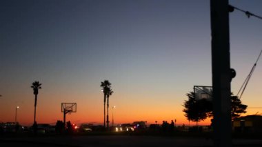 Basketbol sahasındaki oyuncuların siluetleri, basketbol oynayan insanlar, günbatımı okyanus plajı, Kaliforniya sahili, ABD. Sokak sporu sahasında siyah halka, ağ ve arkabahta. Görev plajı.